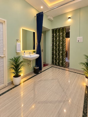 A bathroom with a sink, mirror, and blue curtain. There are potted plants and a trash bin. - Geeta Bhawan Cozy Homestay (Varanasi)