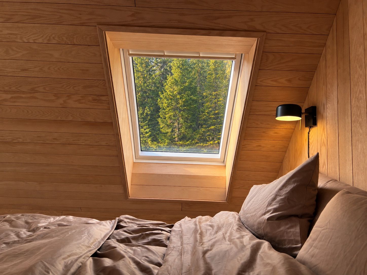 Un dormitorio con tragaluz, paredes de madera, una cama con almohadas y una lámpara fijada a la pared.