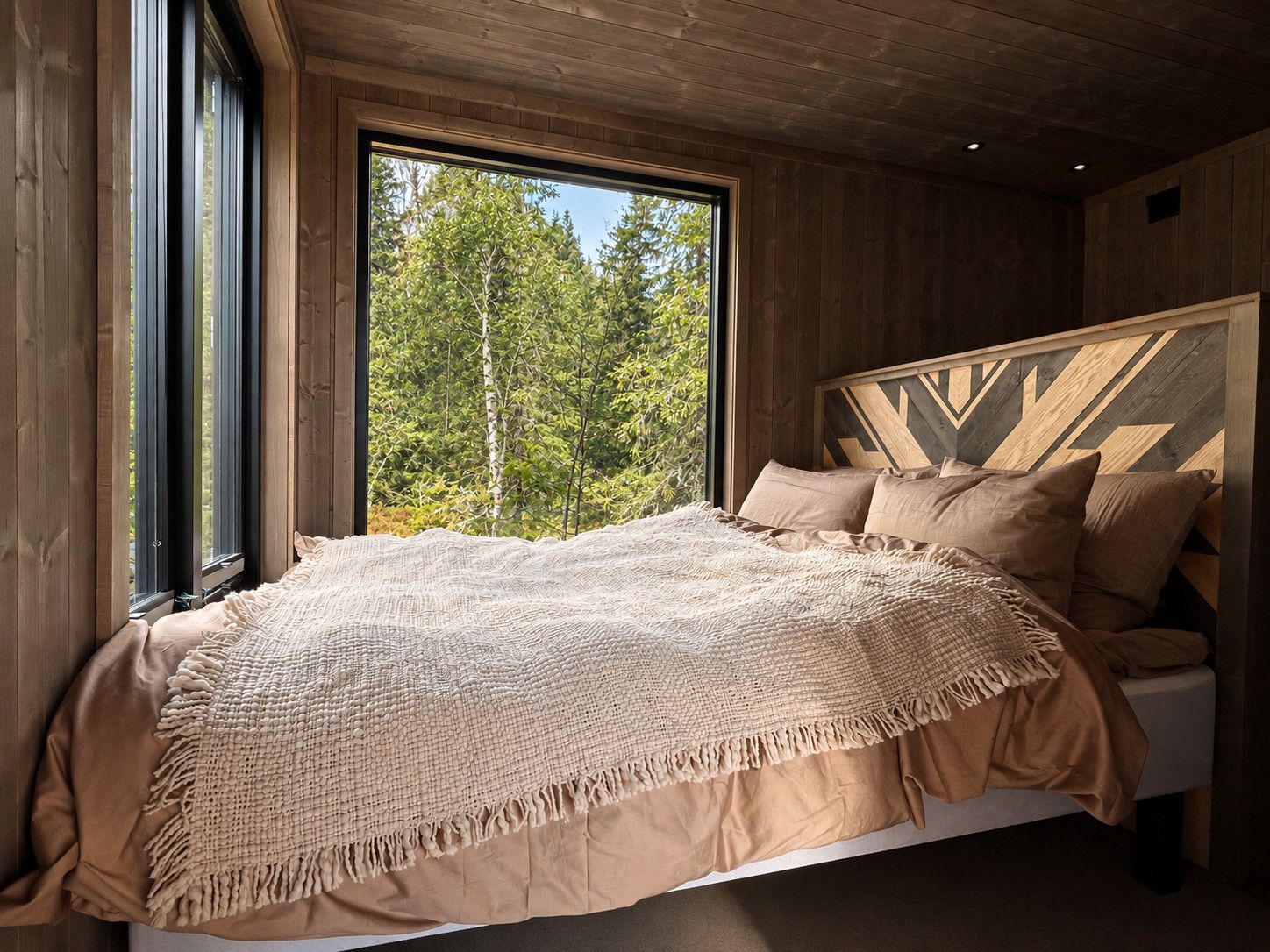 A bedroom with a large window, a bed with a patterned blanket, and a wooden headboard.