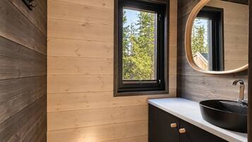 A modern bathroom with wooden panel walls, a window with a view of trees, a round mirror, and a black sink with a white countertop.