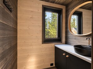 A modern bathroom with wooden panel walls, a window with a view of trees, a round mirror, and a black sink with a white countertop. - Modern and Idyllic Mountain Retreat in Hemsedal (Hemsedal)