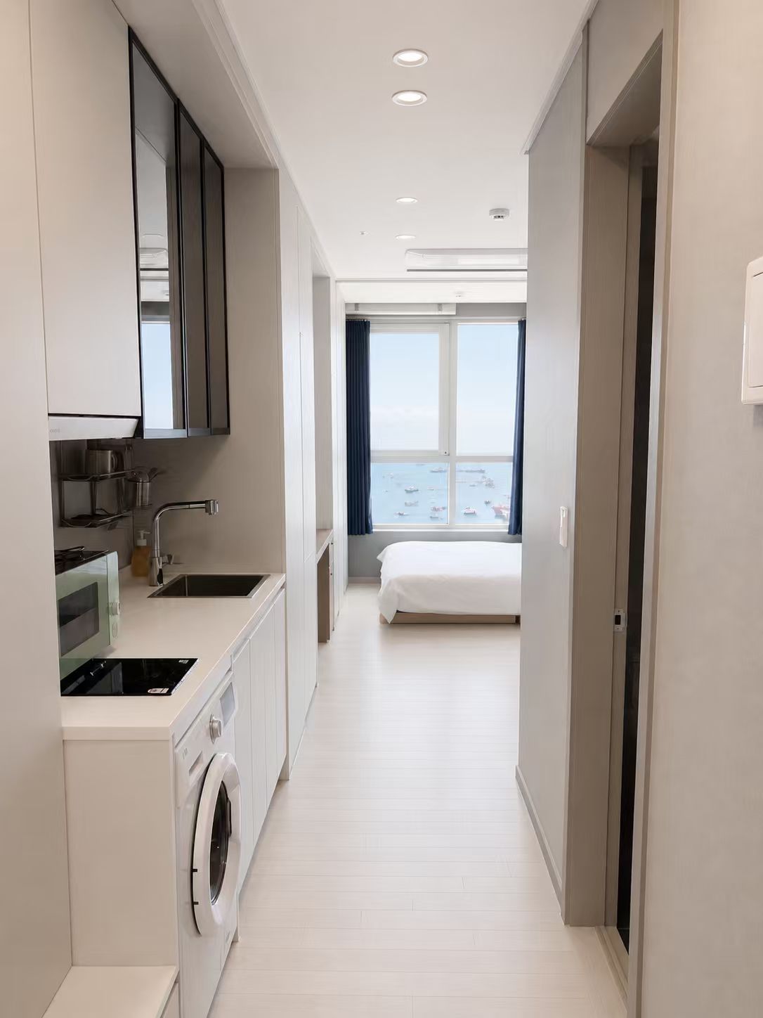 A modern kitchen with a washing machine, a bed, and a view of the sea.