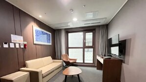 Habitación de hotel con sofá, sillón, mesita, televisor y ventana con cortinas.