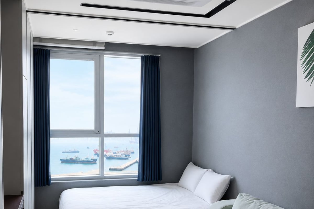 A modern hotel room with a large bed, a sofa, a view of the sea, and a ceiling air conditioning unit.