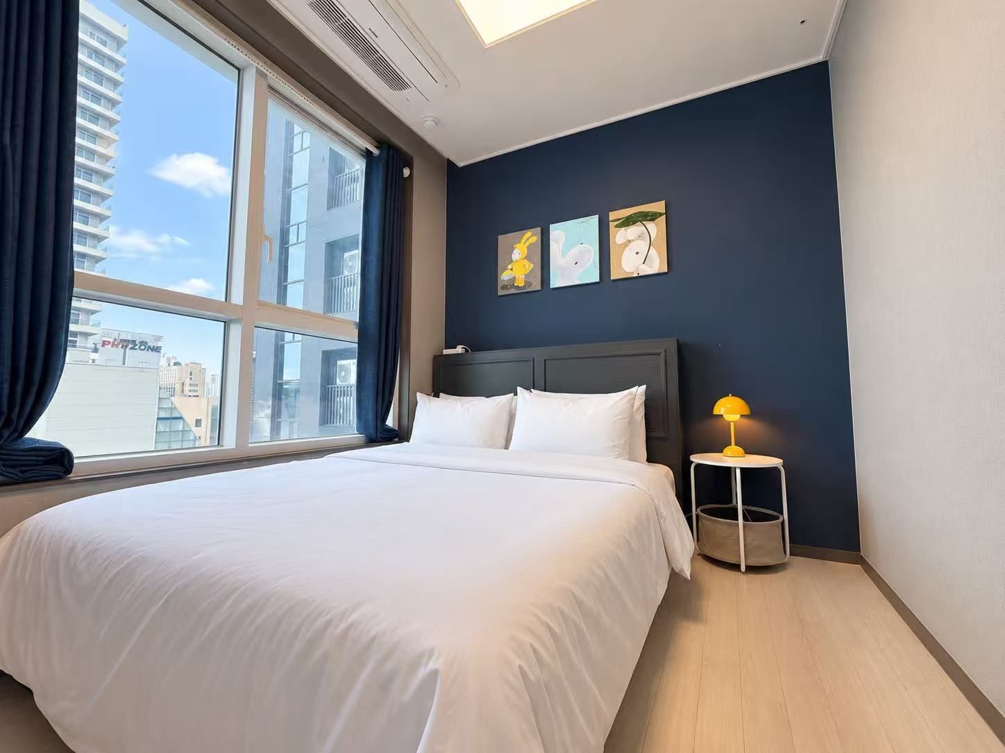 A hotel room with a large bed, a nightstand with a lamp, and a view of the city through a large window.