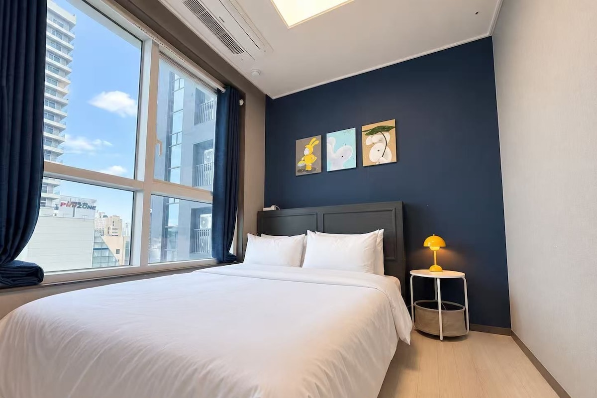 A hotel room with a large bed, a nightstand with a lamp, and a view of the city through a large window.