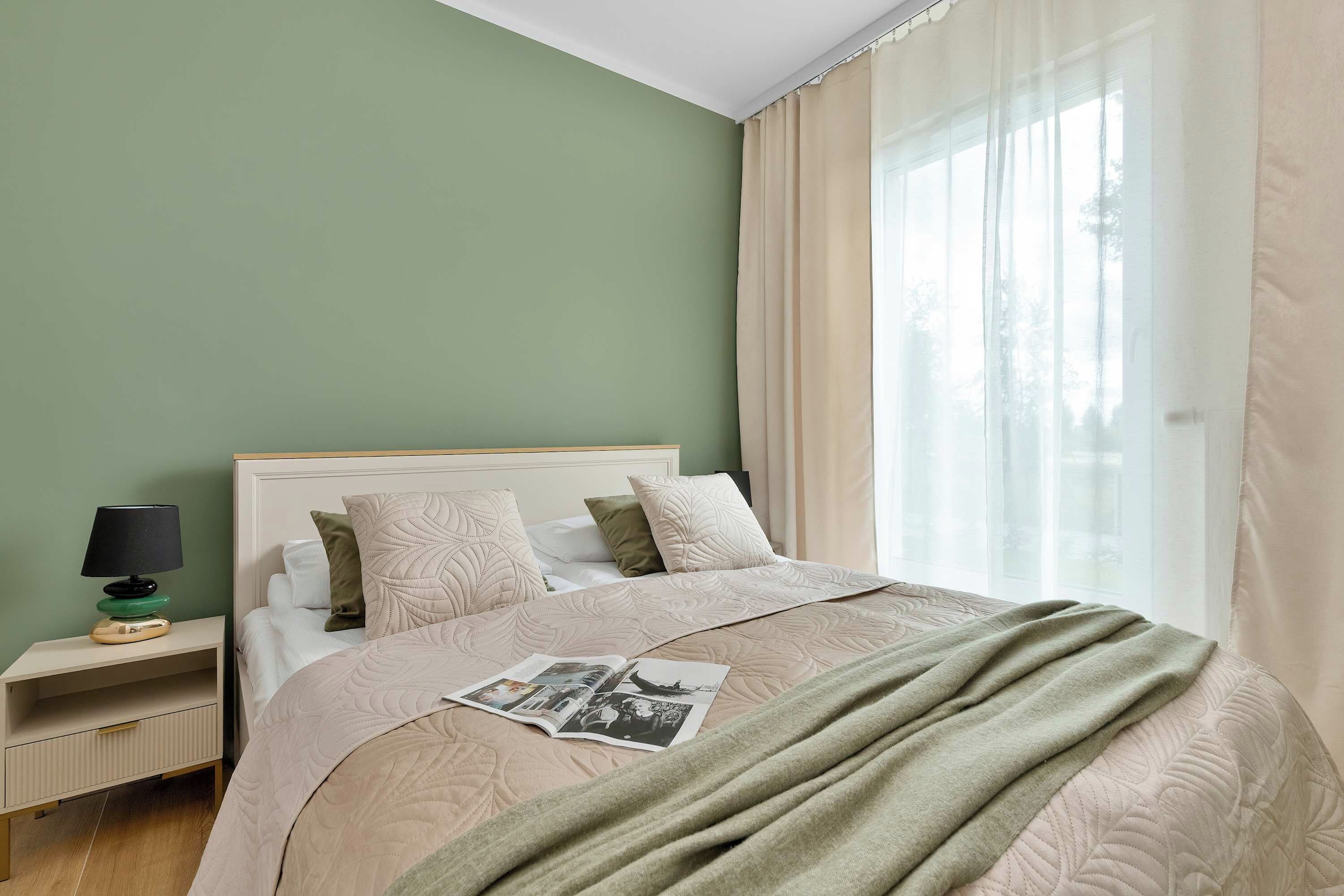 Cama bem arrumada com colcha, travesseiros e uma revista. Um criado-mudo com luminária e um livro. O quarto tem uma parede verde e uma janela com cortinas.