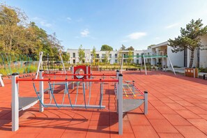 Un patio de recreo al aire libre con diversos juegos, rodeado de una zona pavimentada y edificios al fondo.