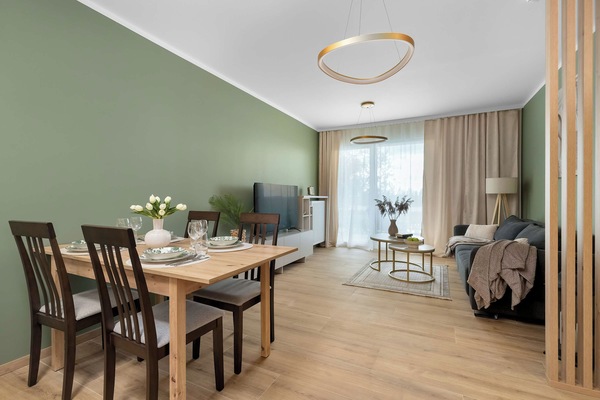 A bright dining and kitchen area with a wooden dining table and chairs. The open-plan space features light furnishings, a kitchen area, and large windows that let in plenty of natural light.
