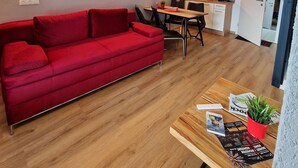 Et moderne opholdsrum med en rød sofa, trægulv, et spiseområde med bord og stole, og et køkken i baggrunden.