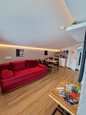 Een moderne woonruimte met een rode bank, houten vloeren, een eethoek met tafel en stoelen, en een keuken op de achtergrond.