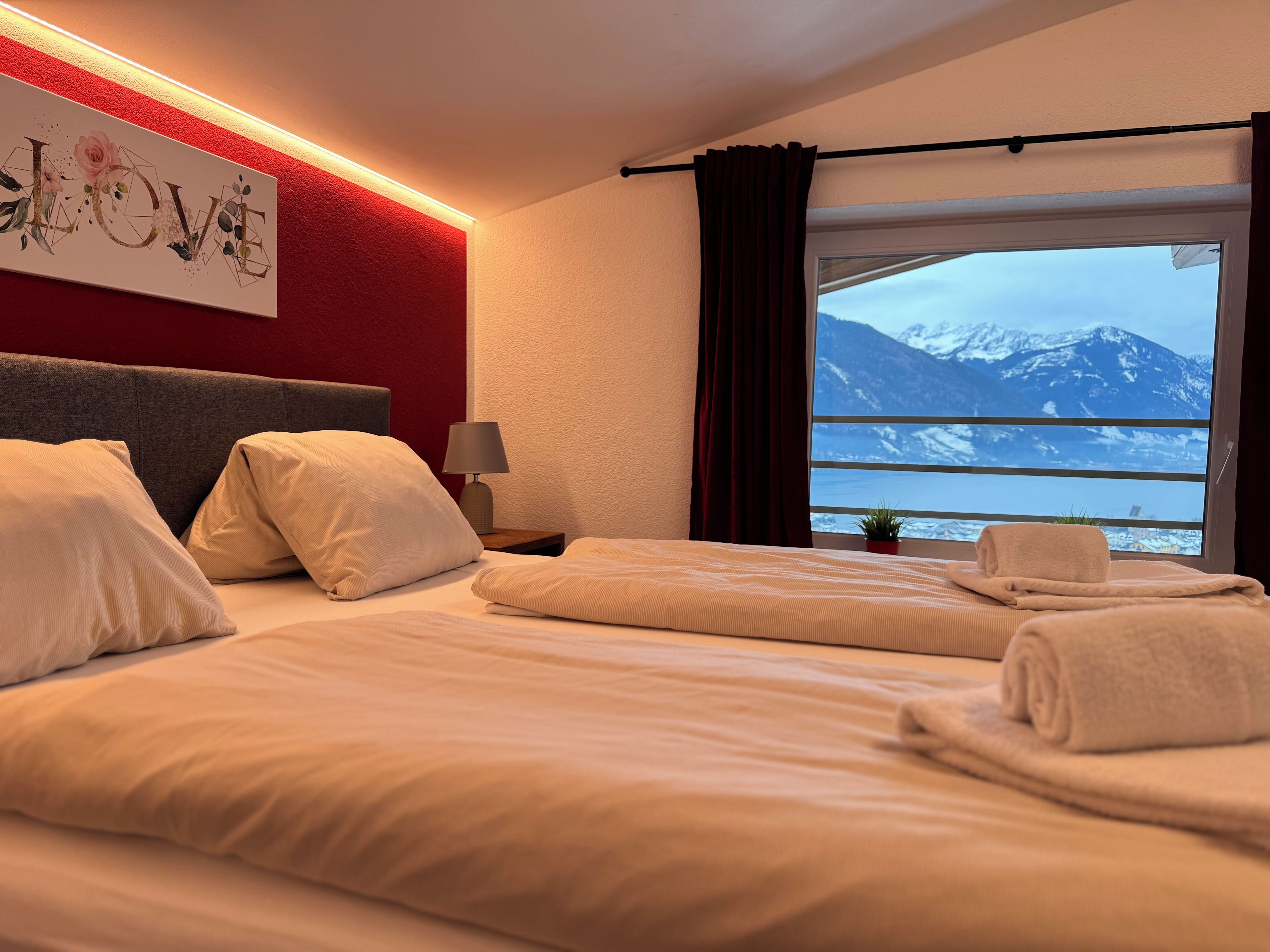 A hotel room with a bed, a nightstand, a lamp, a window with a view of mountains, and a wall with a 'LOVE' sign.