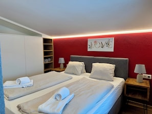 Een hotelkamer met twee bedden, een rode accentmuur, kunstwerk met de tekst 'LOVE' en netjes gevouwen handdoeken.