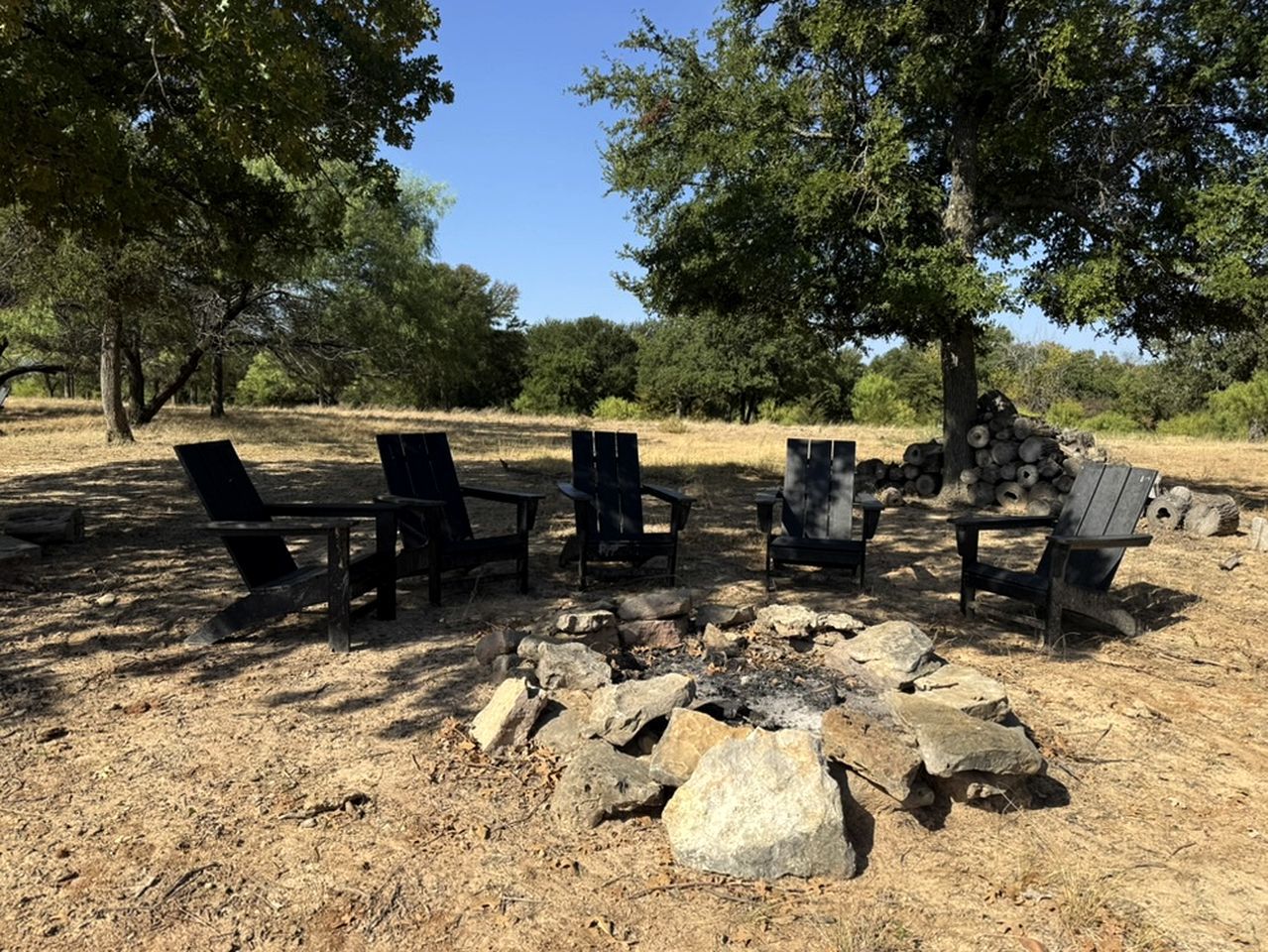 Un terrain de camping avec un foyer entouré de chaises et de tables, aménagé dans une zone herbeuse et aride, avec des arbres en arrière-plan.