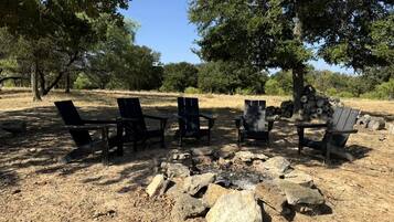Un terrain de camping avec un foyer entouré de chaises et de tables, aménagé dans une zone herbeuse et aride, avec des arbres en arrière-plan.
