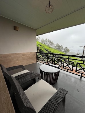 A covered outdoor seating area with wicker furniture and a table, overlooking a hilly landscape.