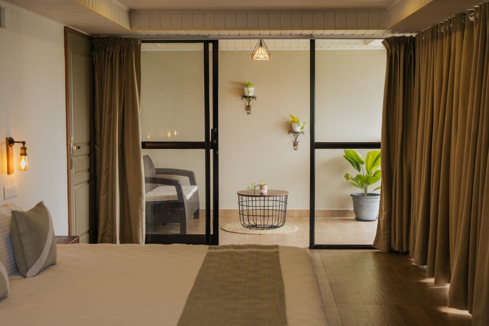 Habitación de hotel con una cama, una mesita, una silla, una planta en maceta y un ventanal corredizo que da a un espacio exterior.