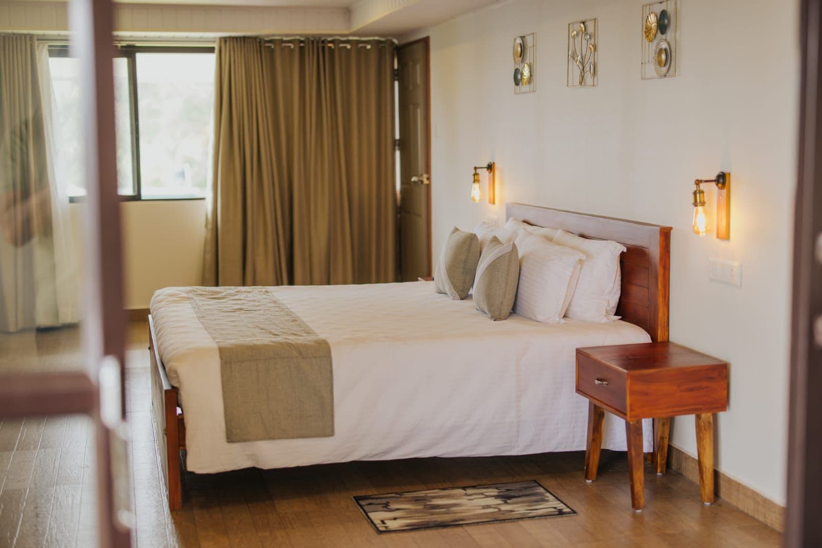 A hotel room with a large bed, a wooden bedside table, and a nightstand. There are wall sconces on either side of the bed.