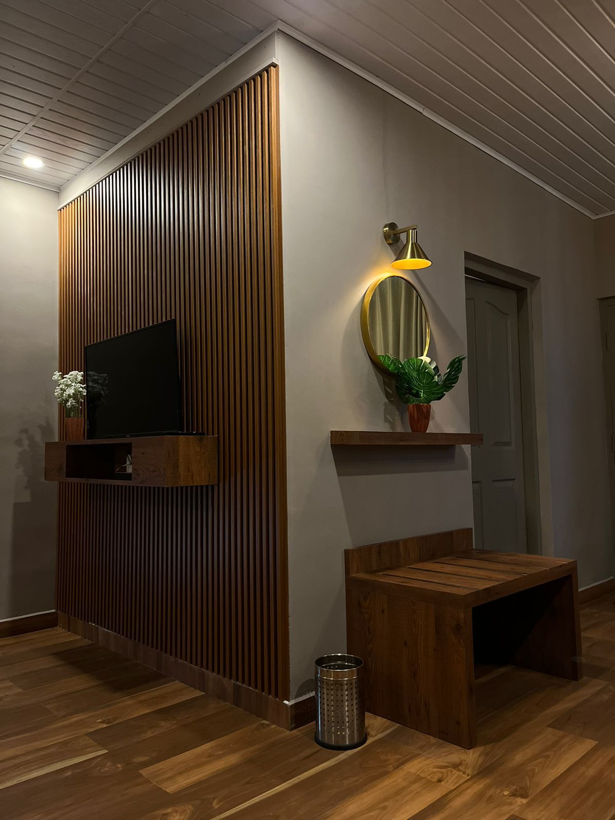 Una sala moderna con una pared de acento de madera, un televisor montado, una lámpara de pared, un banco de madera y un cubo de basura.