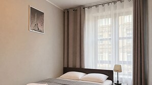 A hotel room with a bed, a nightstand with a lamp, a window with curtains, and a framed picture on the wall. - MARGI City 2B Apartament (Szczecin)