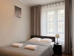 Una habitación de hotel con cama, una mesita de noche con lámpara, una ventana con cortinas y un cuadro en la pared.