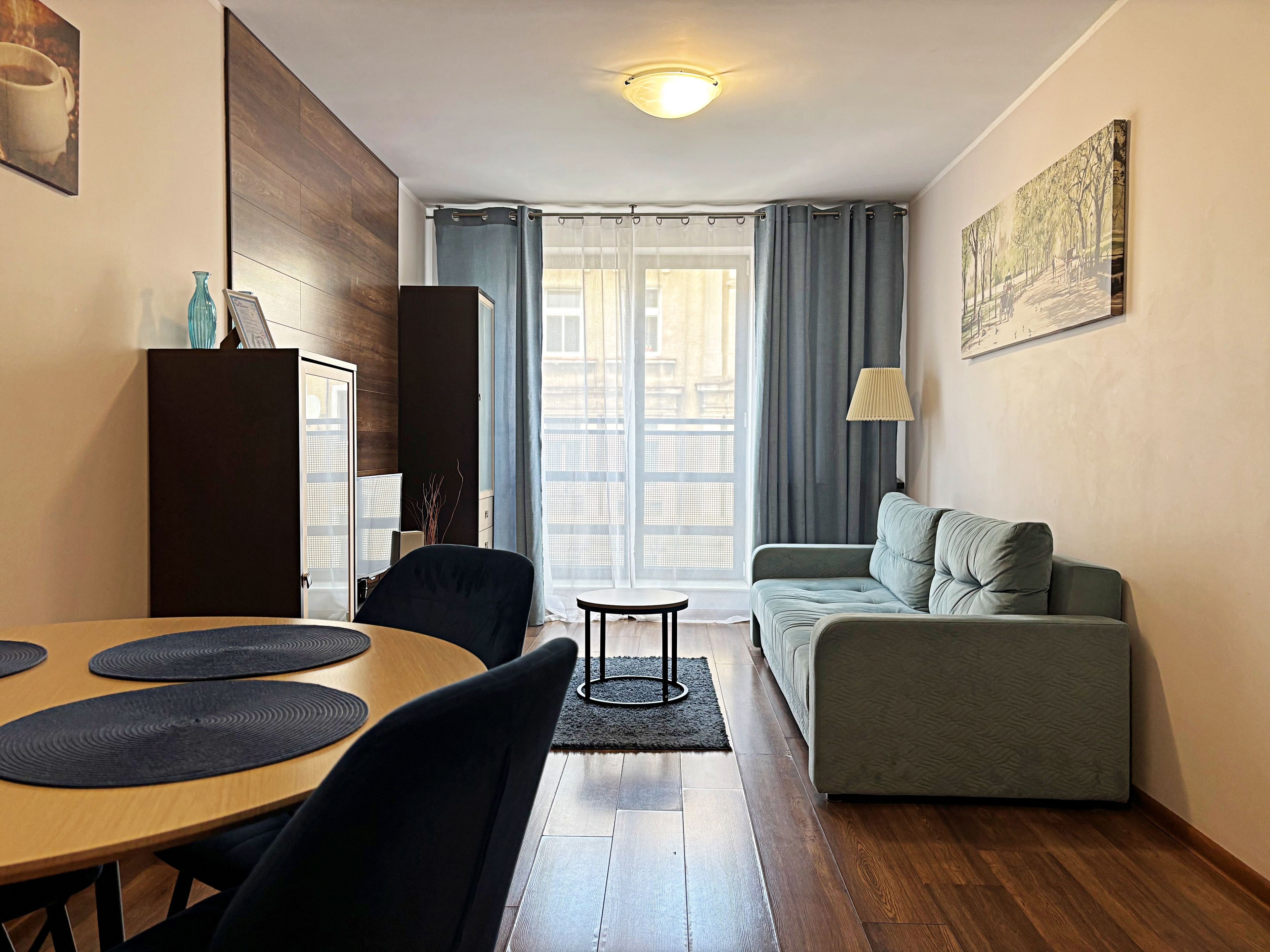 Ein modernes Wohnzimmer mit Holzboden, einem grauen Sofa, einem Esstisch mit schwarzen Platzsets, einem schwarzen Stuhl, einem kleinen runden Tisch und einer Lampe.