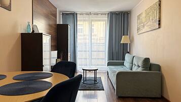 Ein modernes Wohnzimmer mit Holzboden, einem grauen Sofa, einem Esstisch mit schwarzen Platzsets, einem schwarzen Stuhl, einem kleinen runden Tisch und einer Lampe.
