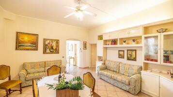 A living room with two sofas, a dining table with chairs, a ceiling fan, and a kitchen area with cabinets and shelves.