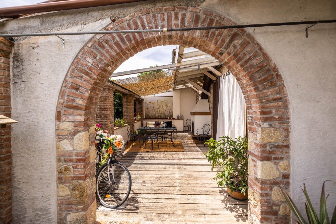 An outdoor patio area with a brick archway, wooden flooring, a table with chairs, and potted plants.