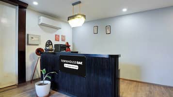 A reception desk with a sign indicating it is company-serviced, a potted plant, a ceiling light fixture, and framed artwork on the wall.