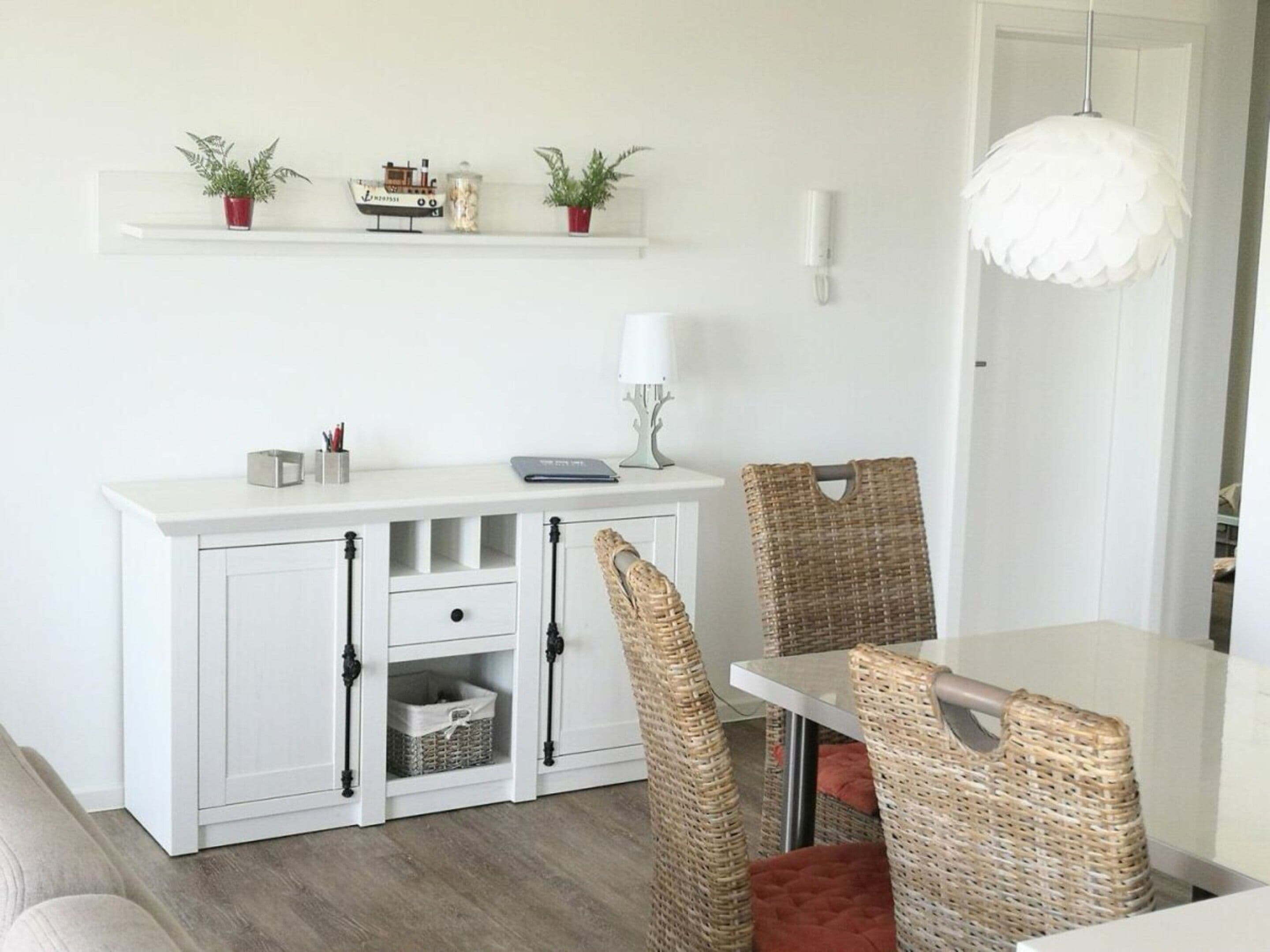 Uma área de refeições com mesa branca, cadeiras de vime, armário branco com gavetas e luminária pendente.
