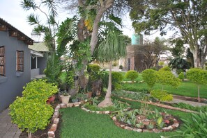 Um jardim bem cuidado com diversas plantas e uma árvore, cercado por um muro de tijolos e um edifício com fachada cinzenta.