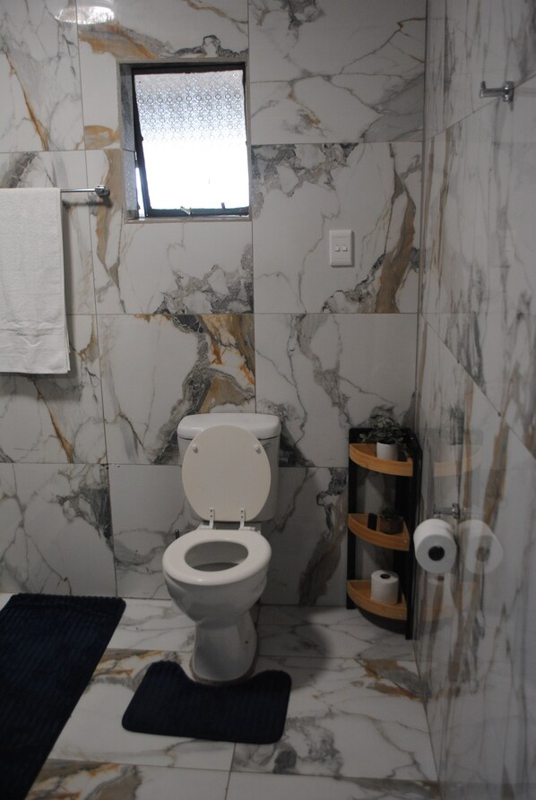Casa de banho com sanita, uma pequena janela, um porta-toalhas e uma prateleira com artigos de higiene pessoal.