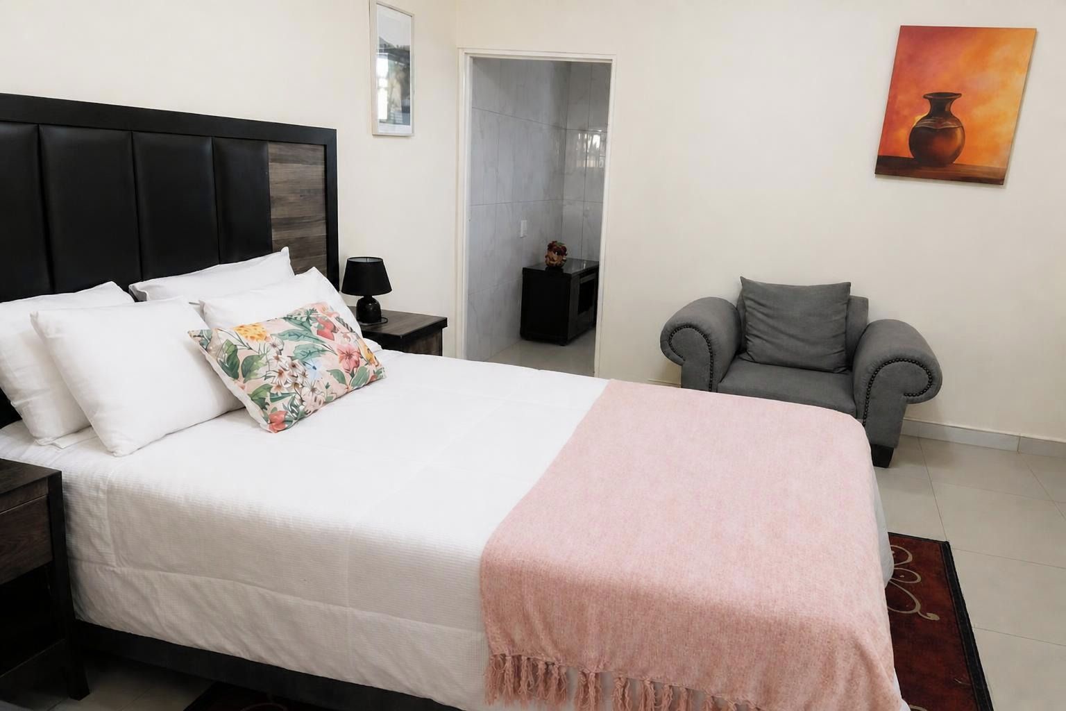 Un lit bien fait, avec du linge de lit blanc et un coussin à motifs floraux, un fauteuil gris et un tableau accroché au mur.