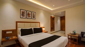 Une chambre d’hôtel avec un grand lit, des tables de chevet, un bureau et une chaise. Des tableaux encadrés ornent le mur, et une porte donne accès à une autre pièce.