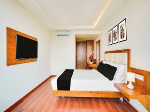 Habitación de hotel con cama, televisor montado en la pared, cabecero de madera y mesita de noche con lámpara.