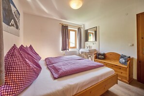 Ein Schlafzimmer mit einem Bett, einer hölzernen Bank, einem Fenster mit Vorhängen, einem Spiegel und einem Badezimmer mit einem Waschbecken.