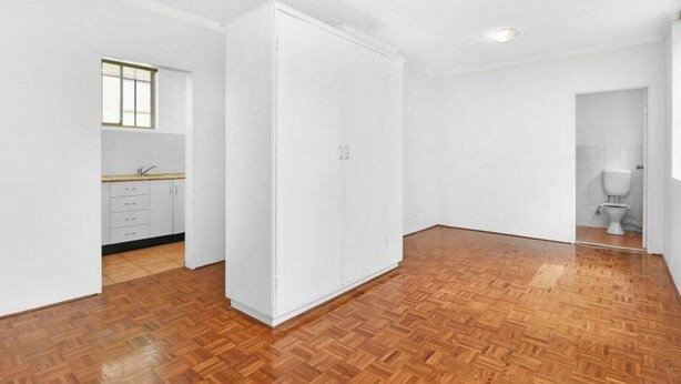 Una habitación con piso de parquet de madera, paredes blancas, una cocina con armarios y un ventanal, y una puerta que da a un baño con inodoro.