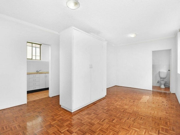 Een kamer met een houten parketvloer, witte wanden, een keuken met keukenkastjes en een raam, en een deur die leidt naar een badkamer met een toilet.