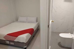 Una cama individual con una colcha roja, un almohadón blanco y cabecera blanca en una habitación con suelo de mármol y un inodoro blanco.