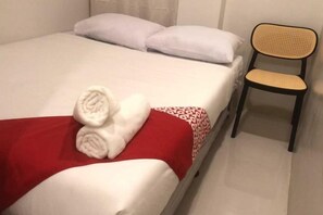 Una cama con ropa blanca, una manta roja y toallas enrolladas. Hay una silla con respaldo tejido.