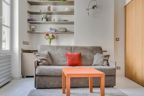 Ein modernes Wohnzimmer mit einem grauen Sofa, einem kleinen orangefarbenen Tisch und einer an der Wand befestigten Regal mit verschiedenen Gegenständen.