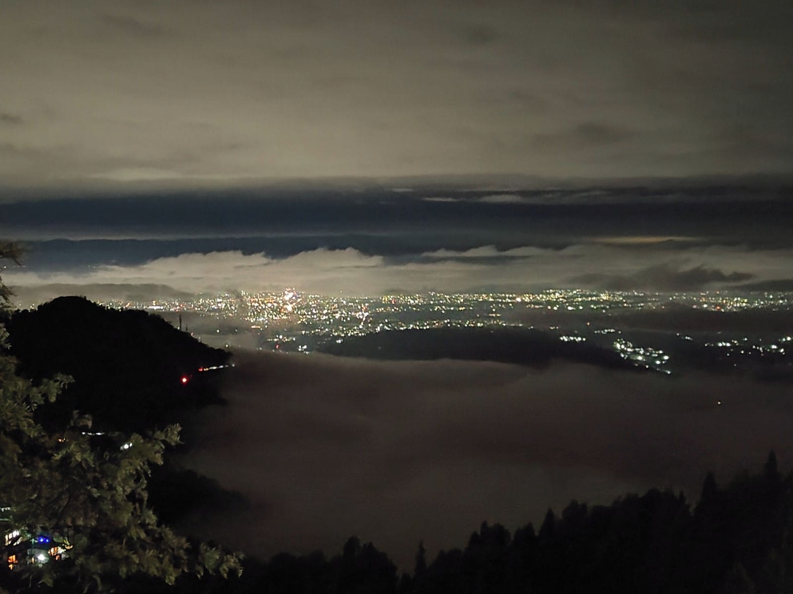 A nighttime cityscape with a view of the city lights below a cloud-covered mountain range.