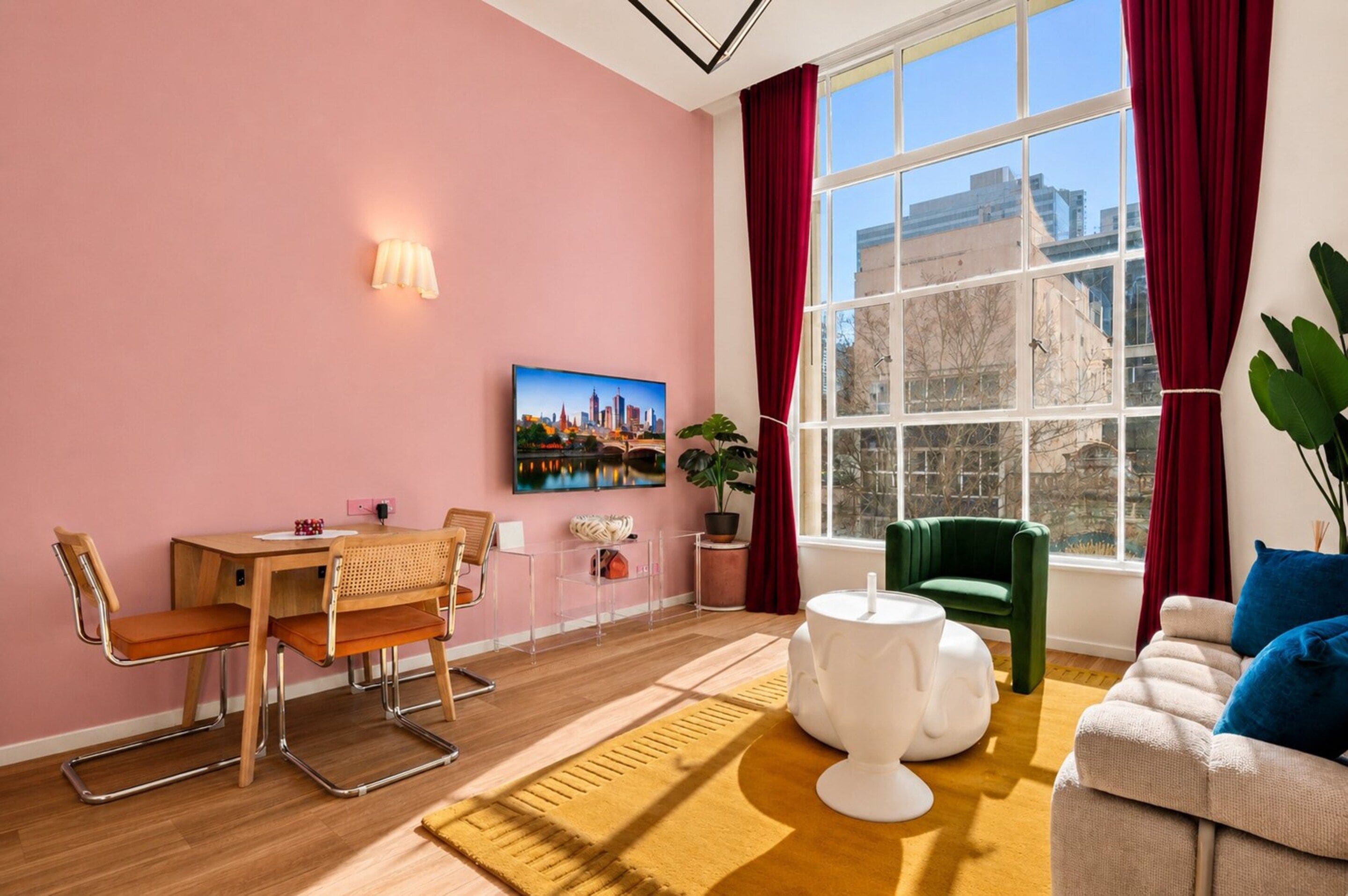 A room with a wooden dining table, two chairs, a green sofa, a white coffee table, and a large window with red curtains.