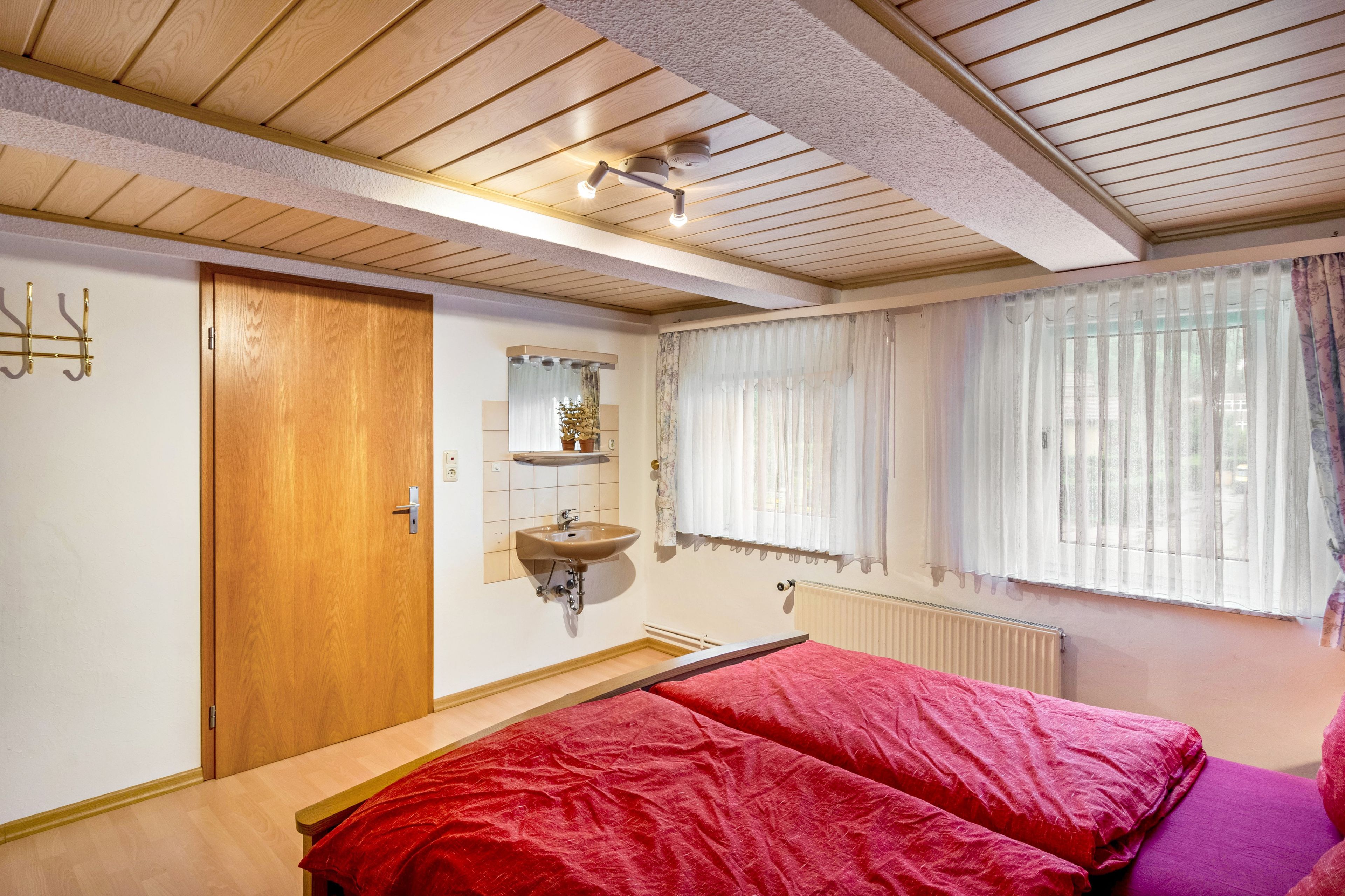 A bedroom with a wooden ceiling, a bed with red bedding, a bedside table, a window with sheer curtains, and a radiator.