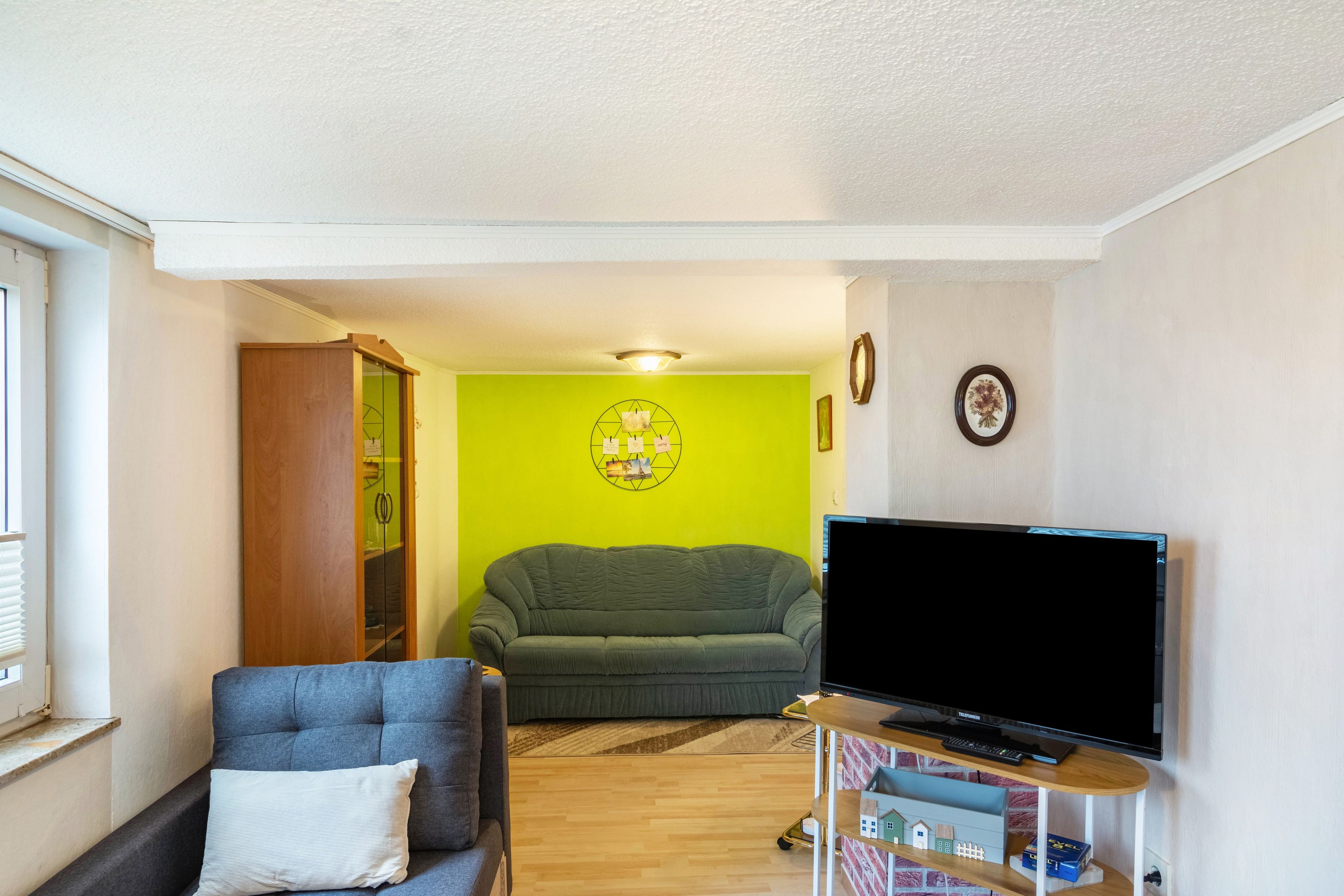 Ein Wohnzimmer mit grüner Wand, einem grauen Sofa, einem blauen Sessel, einem Fernseher auf einem Standfuß und einem kleinen Tisch mit einer Tastatur.