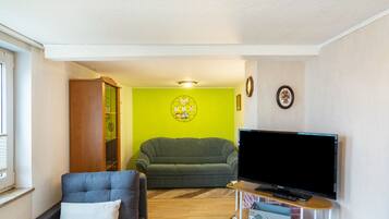 Ein Wohnzimmer mit grüner Wand, einem grauen Sofa, einem blauen Sessel, einem Fernseher auf einem Standfuß und einem kleinen Tisch mit einer Tastatur.