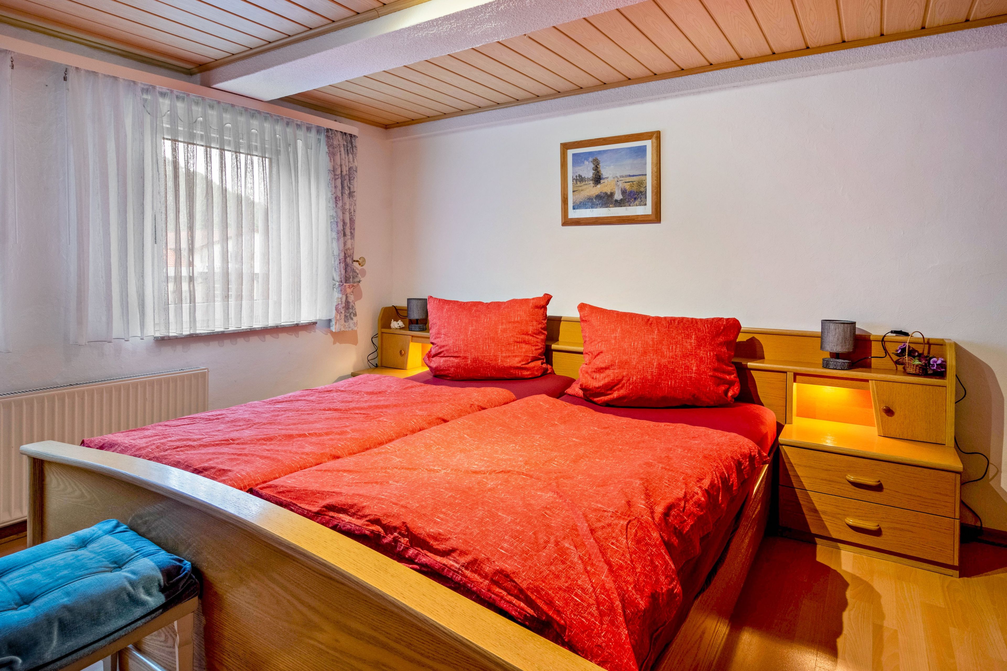 Een slaapkamer met een houten bed, rode beddengoed, een nachtkastje met een lamp en een raam met dunne gordijnen.