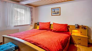 Een slaapkamer met een houten bed, rode beddengoed, een nachtkastje met een lamp en een raam met dunne gordijnen.