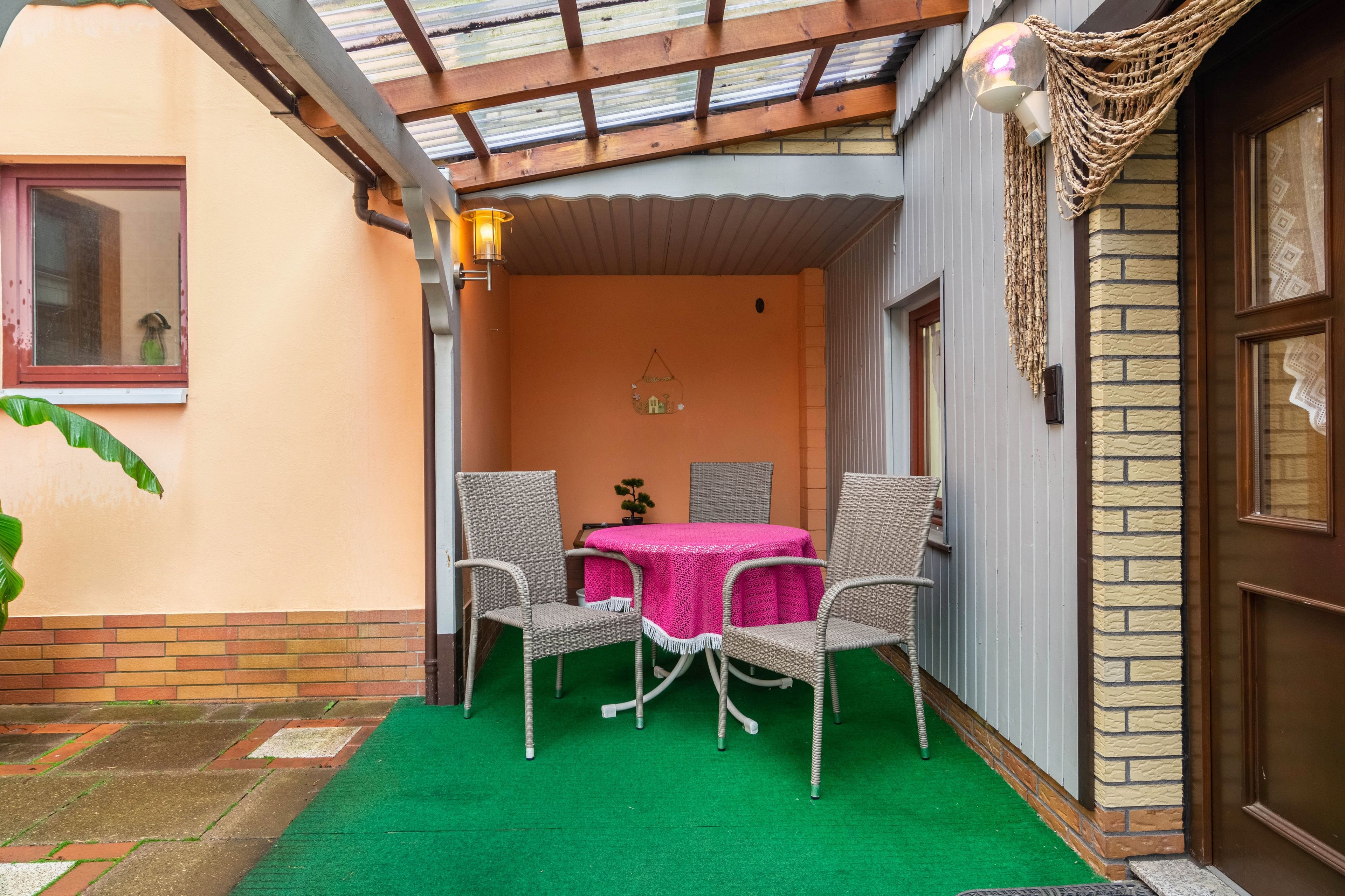 Een overdekt terras met een tafel, stoelen, een deur en een muur met een decoratieve wandhanger.